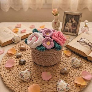 Puede incluir: Un arreglo floral de ganchillo en una cesta tejida, con flores rosas, blancas y moradas. El arreglo está sobre un salvamanteles tejido, rodeado de pétalos esparcidos y adornos en forma de corazón. También hay una foto enmarcada y libros abiertos.