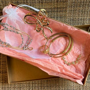 May include: A collection of gold-toned jewelry and accessories including a butterfly-shaped hair claw, a delicate star-patterned necklace, two textured bangles, a ring, and a multi-strand bracelet with small grey beads, all presented in a gift box.