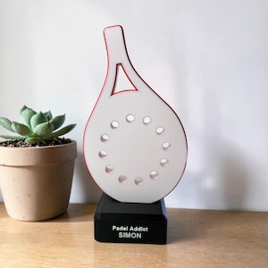 Peut inclure: Trophée en forme de raquette de padel blanche et rouge, sur une base noire. Le trophée présente des découpes circulaires et le texte "Padel Addict SIMON". Un petit pot de plante succulente est à gauche.