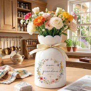 May include: A white ceramic vase with a scalloped top, filled with colorful flowers. The vase has a floral design and the words "mom grandma" with dates. A gold ribbon is tied around the vase.