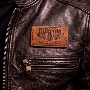 K&ouml;nnte beinhalten: Nahaufnahme einer braunen Lederjacke mit einem Aufn&auml;her mit der Aufschrift "AMERICAN CUSTOM" &uuml;ber einer Illustration von Fl&uuml;geln und einer Z&uuml;ndkerze. Der Text "RIDE FAST OR DIE LAST" steht darunter. Die Jacke hat eine Rei&szlig;verschlusstasche.
