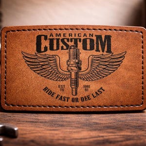 Puede incluir: Parche rectangular de cuero marrón con las palabras "AMERICAN CUSTOM" en negro. Presenta un diseño de bujía alada y el texto "RIDE FAST OR DIE LAST". El fondo está borroso.