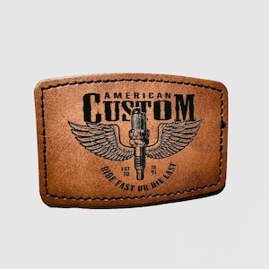 Peut inclure: Un patch rectangulaire en cuir marron avec des coutures noires. Il affiche les mots "AMERICAN CUSTOM" au-dessus d'un motif de bougie d'allumage ailée. En dessous, on lit "RIDE FAST OR DIE LAST" et les années "EST 10" et "19 91".