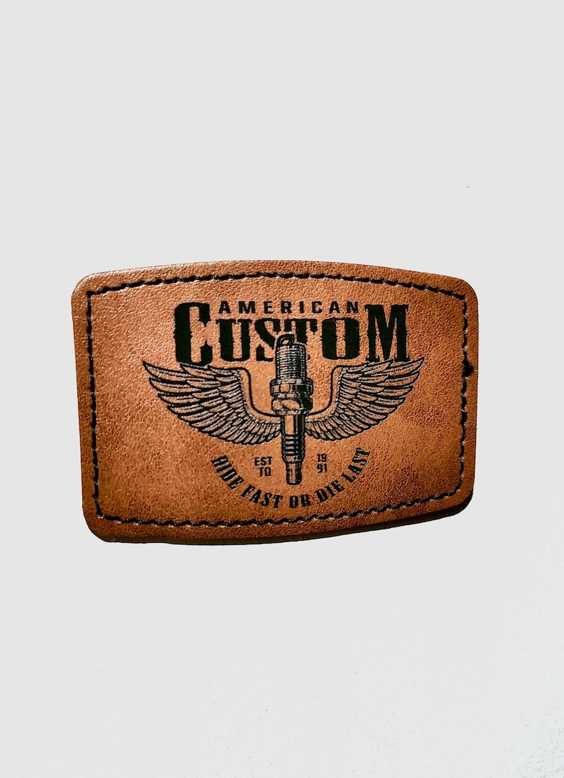 K&ouml;nnte beinhalten: Ein brauner Lederaufn&auml;her mit schwarzen N&auml;hten. Der Aufn&auml;her zeigt die Worte "AMERICAN CUSTOM" &uuml;ber einer Grafik einer Z&uuml;ndkerze mit Fl&uuml;geln. Darunter steht der Text "RIDE FAST OR DIE LAST" und die Jahreszahlen "EST 10" und "19 91".