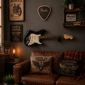 Peut inclure: Une guitare électrique noire avec un pickguard blanc fixée sur un mur sombre au-dessus d'un canapé en cuir marron. Le mur présente un panneau en forme de médiator Fender, une affiche de moto et un panneau Dad's Garage. Deux coussins décoratifs à motifs de moto reposent sur le canapé.