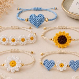 Op de afbeelding: Een verzameling handgemaakte macramé-armbanden. De armbanden hebben verschillende ontwerpen, waaronder een blauw hart, madeliefjes, een zonnebloem en een combinatie van harten en madeliefjes. Elke armband heeft goudkleurige kralen en verstelbare koorden.