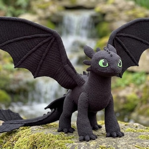 May include: A black crocheted dragon with large wings and green eyes. The dragon is standing on a mossy surface with a waterfall in the background. The wings are spread, and the tail is long and curved.