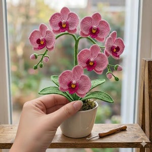 May include: A handcrafted crochet orchid plant in a white pot. The pink flowers have dark pink centers and yellow accents. The green leaves and stem add contrast. A crochet hook rests on a wooden shelf.