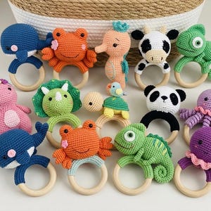 May include: A collection of handmade crochet animal rattles featuring a blue whale, orange crab, peach seahorse, black and white cow, green chameleon, pink dinosaur, green triceratops, turtle, panda, and purple octopus, each attached to a smooth wooden ring.