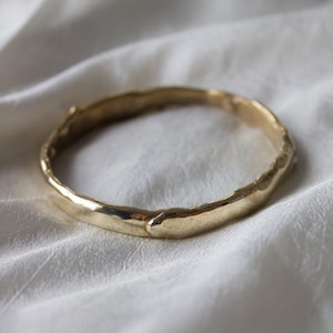 May include: A gold-colored bangle bracelet with a textured surface. The bracelet is circular and appears to be made of metal. The background is a white, silky fabric.