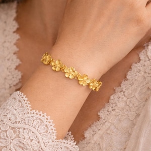 Peut inclure: Un bracelet doré orné d'un motif floral. Le bracelet est composé de maillons en forme de fleurs, créant un accessoire délicat et élégant. Il est porté au poignet, mettant en valeur ses détails et son savoir-faire.