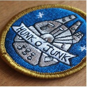 May include: A blue and gold embroidered iron-on patch featuring a stylised image of the Millennium Falcon with the text "Hunk o' Junk 888" on a banner.