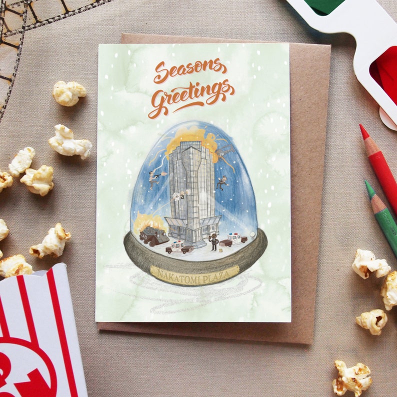 May include: A Christmas card featuring a snow globe with a stylized illustration of the Nakatomi Plaza building from the movie Die Hard. The snow globe is surrounded by a light green background with white snowflakes. The text "Seasons Greetings" is written in brown cursive at the top of the card.