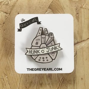 May include: A wooden pin shaped like a spaceship with a banner across the centre that reads HUNK O' JUNK. The pin is mounted on a white square display card featuring the text The GREY EARL and THEGREYEARL.COM in black ink.