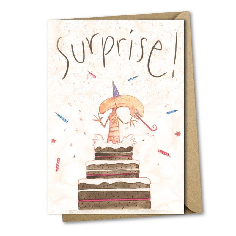 May include: A humorous birthday card featuring a cartoon alien wearing a party hat and popping out of a layered chocolate cake. The card reads "Surprise!" in a whimsical font.