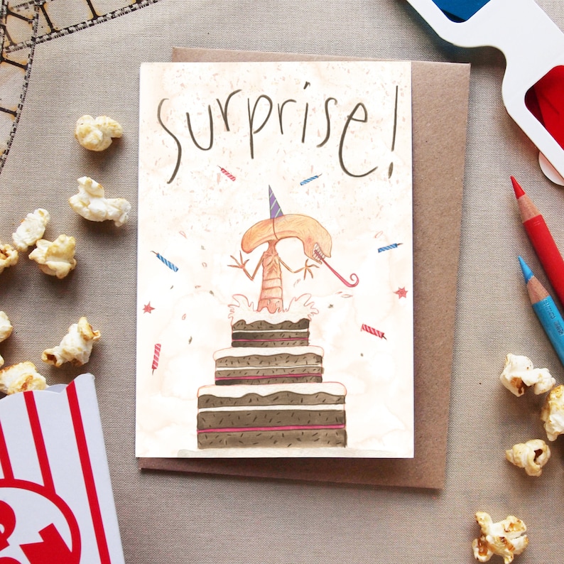 May include: A birthday card with a watercolor illustration of a creature jumping out of a chocolate cake. The card has the text "Surprise!" written in a whimsical font.