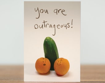 May include: A greeting card with the text "you are outrageous!" and a drawing of a cucumber and two oranges with faces.