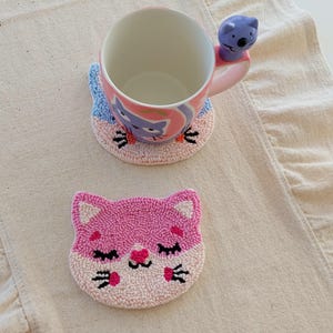 May include: A pink and white cat-themed mug and matching coaster set. The mug features a purple cat design and a blue koala handle. The coaster is a pink cat face with black whiskers and closed eyes. The set is on a beige surface.