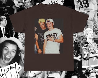 May include: A dark brown short-sleeve t-shirt featuring a central rectangular graphic print of two individuals wearing baseball caps. One individual wears a black t-shirt with white text, and the other wears a white t-shirt with the text CRAZY KIDS.