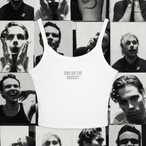 May include: A white sleeveless crop top featuring a scoop neckline and thin shoulder straps. The center of the chest displays the text DIBS ON THE BASSIST printed in a simple, dark, sans-serif font against the plain white fabric background.