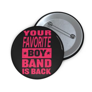 Puede incluir: Dos botones redondos negros con el texto "YOUR FAVORITE BOY BAND IS BACK" en rosa. Un botón está enfocado, el otro es parcialmente visible. Los botones tienen un respaldo de alfiler de metal.