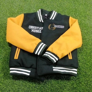 May include: A black and gold varsity jacket featuring white striped ribbed cuffs, collar, and hem. The front displays the embroidered text 'CHRISTIAN MUNOZ' on the right side and a 'G PACKERS' logo on the left side in gold and white thread.