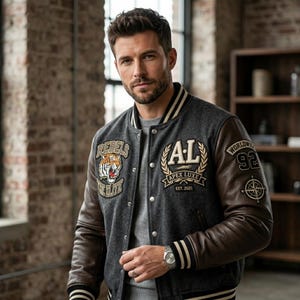 Könnte beinhalten: Eine anthrazitfarbene Collegejacke mit braunen Lederärmeln, verziert mit einem Tiger-Aufnäher, dem Schriftzug Rebels The Elite, einem AL Apex Luxe Est. 2021 Emblem und einem Ärmelaufnäher mit dem Text Worldwide 92 und einem Kompass-Motiv.