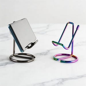 Puede incluir: Dos soportes metálicos para teléfono. Uno es de color gris oscuro, sosteniendo un teléfono. El otro soporte tiene un acabado arcoíris. Ambos soportes tienen una base circular y una cuna curva para sostener un teléfono.