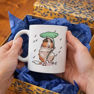 May include: White ceramic mug featuring a cartoon guinea pig holding a leaf umbrella in the rain. The guinea pig is brown, white, and tan. The mug is held in a gold box with blue tissue paper.