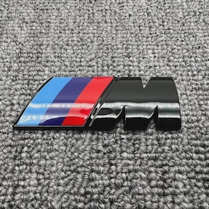Pode incluir: Um emblema preto brilhante e multicolorido com o logótipo BMW M. O emblema tem um design de riscas azuis, azul escuro e vermelhas no lado esquerdo e um "M" preto no lado direito. O emblema está sobre um fundo cinzento texturizado.