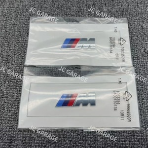 Op de afbeelding: Twee BMW-embleembadges in een doorzichtige plastic verpakking. De badges tonen het BMW-logo in blauw, rood en zilver. De verpakking bevat tekst en nummers. De achtergrond is een grijs oppervlak met textuur.