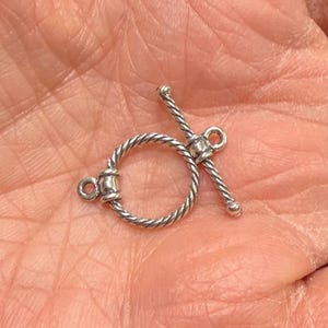 May include: A silver-colored toggle clasp with a twisted rope design. The clasp features a circular ring with a bar that inserts through the ring to secure it. The clasp has two loops for attaching to jewelry.