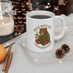 May include: White ceramic coffee mug with the text "Bear With Me I Switched To Decaf" and a cartoon bear illustration. The bear is brown, wearing a green robe, and holding a coffee cup that says "Decaf?".