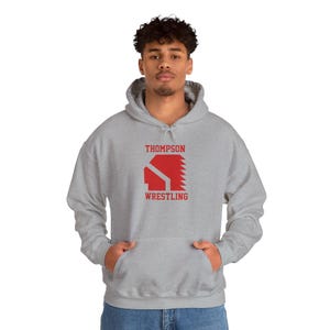 May include: A light gray hooded sweatshirt with a front pocket. The sweatshirt features the text "THOMPSON WRESTLING" in red block letters and a red graphic. The person is wearing blue jeans.