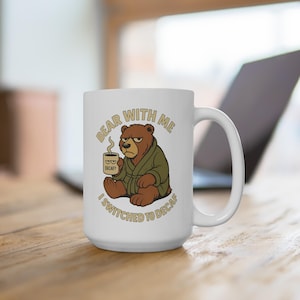 May include: White ceramic mug featuring a cartoon bear in a green robe holding a coffee cup. The mug has the text "BEAR WITH ME I SWITCHED TO DECAF". The bear has a grumpy expression. The mug is on a wooden surface.