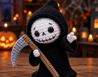 May include: A crocheted grim reaper doll featuring a black hooded robe, a white skull face with black stitched eyes and mouth, pink cheeks, and a small scythe with a grey blade and a brown handle held in one of its white crocheted hands.