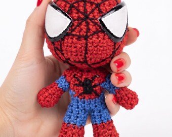May include: A small, handmade crochet doll featuring a red and blue colour scheme. The head is round with a red textured surface, black stitched web patterns, and large white eyes. The body has a blue torso with a black spider emblem and red limbs.