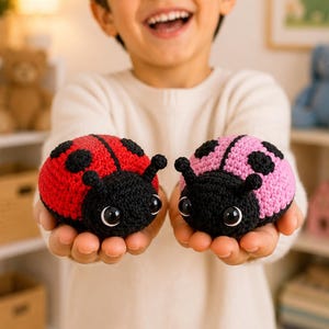 May include: Two small, round, crocheted ladybug toys. One ladybug has a bright red back with black spots, while the other has a soft pink back with black spots. Both feature a black head, two antennae, and large, shiny, round black eyes.