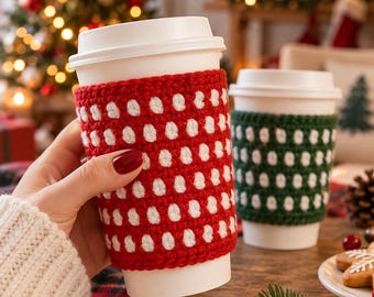 May include: Two white disposable coffee cups with removable crochet sleeves. One sleeve is red with white polka dots, and the other is dark green with white polka dots. The cups feature white plastic lids and are held in a hand.