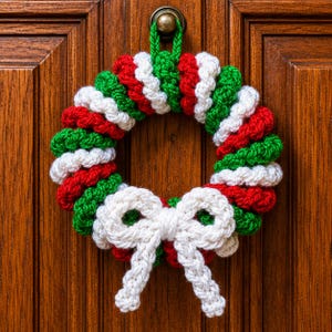 May include: A circular crochet wreath featuring a repeating pattern of red, white, and green yarn segments. The wreath includes a green yarn loop at the top for hanging and a prominent white crocheted bow attached to the bottom center section.