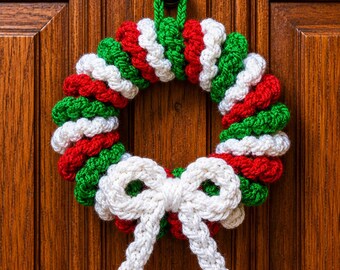 May include: A circular crochet wreath featuring a repeating pattern of red, white, and green yarn segments. The wreath includes a green yarn loop at the top for hanging and a prominent white crocheted bow attached to the bottom centre section.