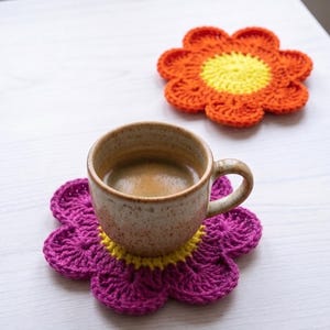 May include: A ceramic mug filled with dark coffee sits on a purple and yellow crocheted flower coaster. A second orange and yellow crocheted flower coaster is positioned nearby. The coasters feature a central yellow circle with petals.