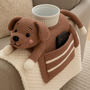 Puede incluir: Organizador de reposabrazos con forma de perro tejido a ganchillo marrón con un soporte para cuenco blanco y un bolsillo. El perro tiene patas blancas y una cola con punta blanca. El bolsillo tiene rayas blancas. Un smartphone negro está en el bolsillo.