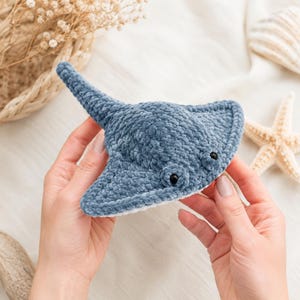 May include: A handmade, blue crochet stingray plush toy with black eyes. The plush has a soft, textured appearance and is held against a backdrop of seashells and dried flowers. A charming, ocean-themed decorative item.