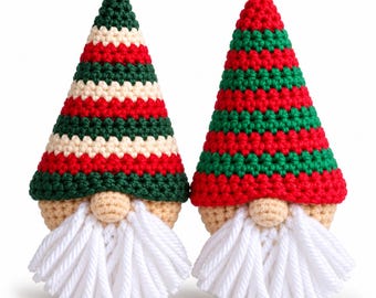 May include: Two crochet gnome ornaments with pointed hats. The left gnome has a green, cream, and red striped hat. The right gnome has a red and green striped hat. Both have beige faces, round noses, and long white yarn beards with hanging loops.