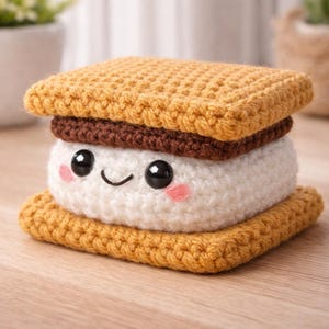 May include: A crocheted s'more toy with a smiling face. The s'more features a white marshmallow centre, a dark brown chocolate layer, and two golden-brown biscuit layers. The marshmallow has black eyes and pink cheeks.