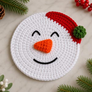 May include: A circular white crochet coaster featuring a snowman face with a black embroidered smile and closed eyes, an orange textured nose, and a red crochet hat brim accented with a small green pom-pom detail on the side.