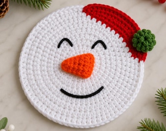 May include: A circular white crochet coaster featuring a snowman face with a black embroidered smile and closed eyes, an orange textured nose, and a red crochet hat brim accented with a small green pom-pom detail on the side.