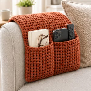 May include: A burnt orange crochet sofa armrest organizer featuring two deep pockets. The left pocket holds a small white book and a pair of brown-rimmed glasses, while the right pocket contains a black smartphone and a black remote control.