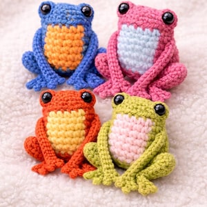 Puede incluir: Cuatro figuras de ranas hechas a mano con ganchillo en azul, rosa, naranja y verde. Cada rana tiene un vientre de color contrastante y ojos de seguridad negros. Las ranas están dispuestas sobre una superficie blanca texturizada.